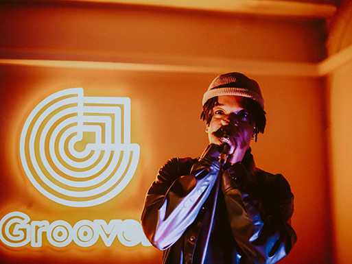 A person sings into a microphone under warm lighting, with a neon 