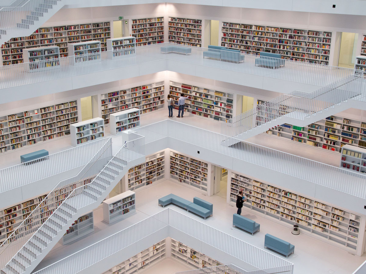 Spacious multi-level library with white walls, bookshelves, blue seating, and people on walkways and stairs.