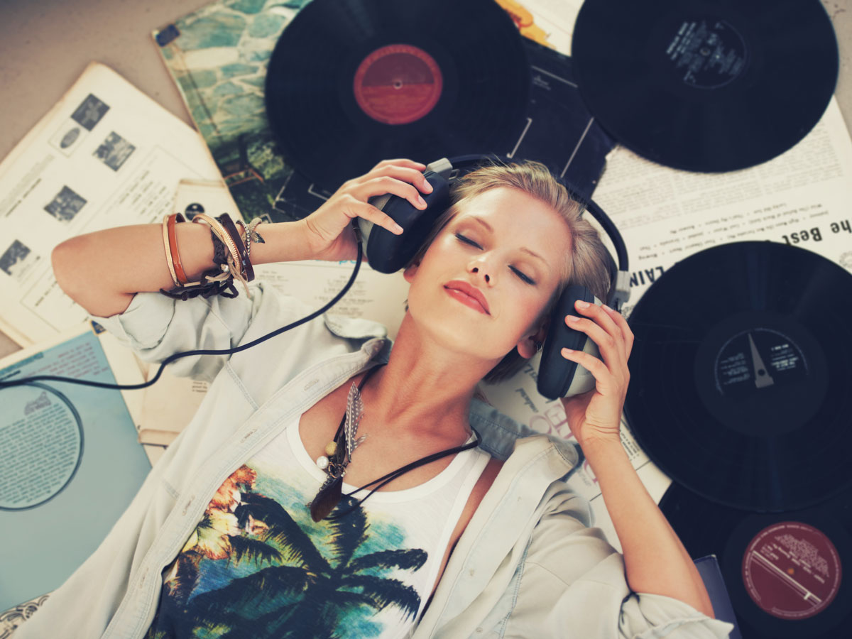 Woman with headphones lies among vinyl records and papers, eyes closed, wearing a palm tree shirt and bracelets.