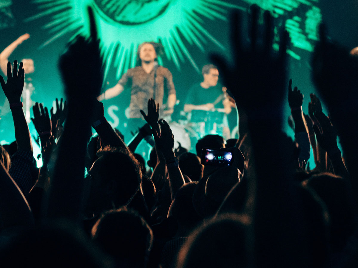 Audience with raised hands at a concert, blurred band on stage with green lights in the background.