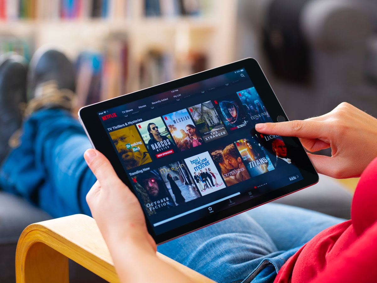 Person in red shirt browsing Netflix on a tablet, selecting shows while lounging on a chair with blurred bookshelves behind.