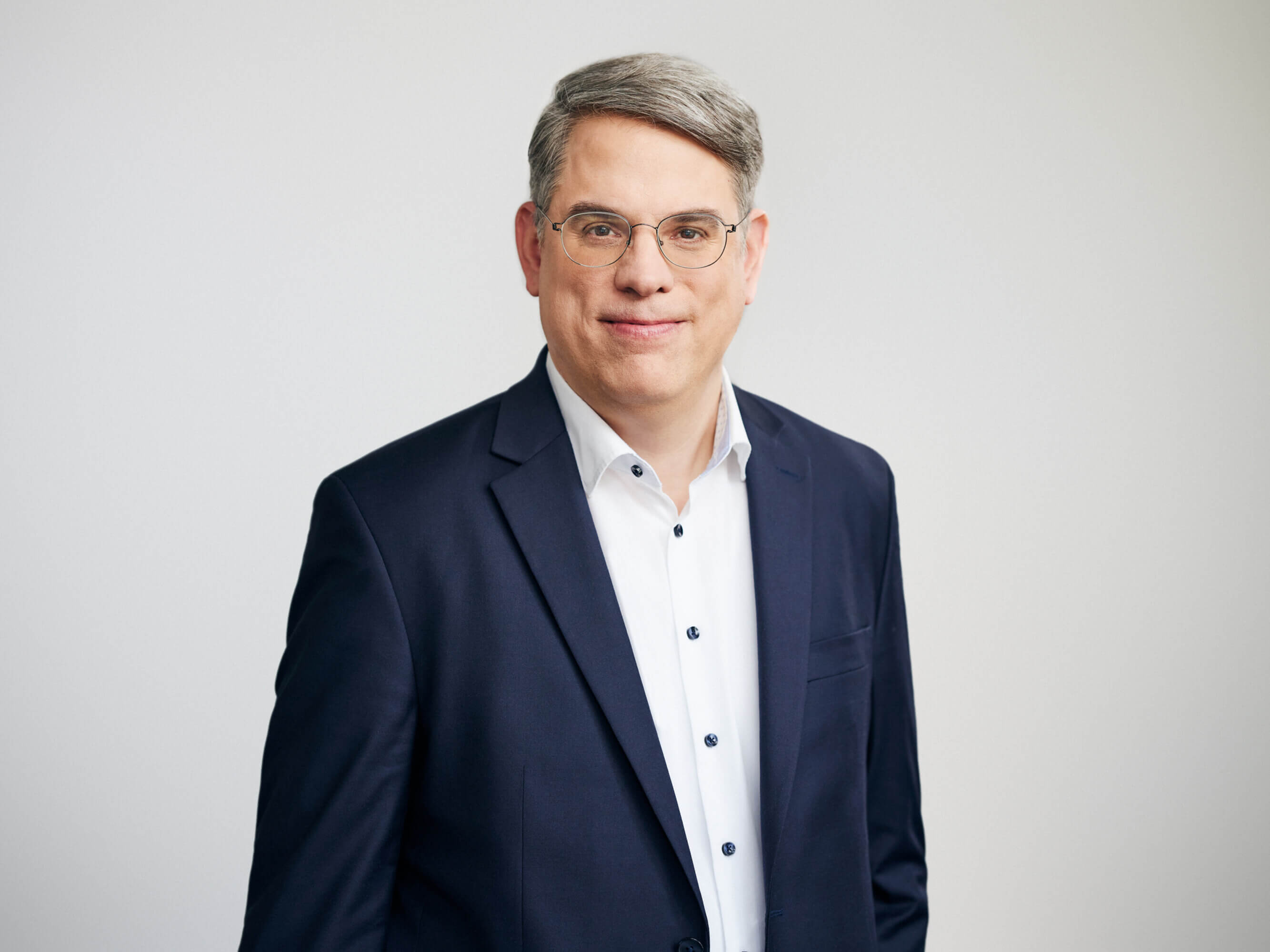 Ein Mann in Anzug und Brille lächelt vor neutralem Hintergrund.