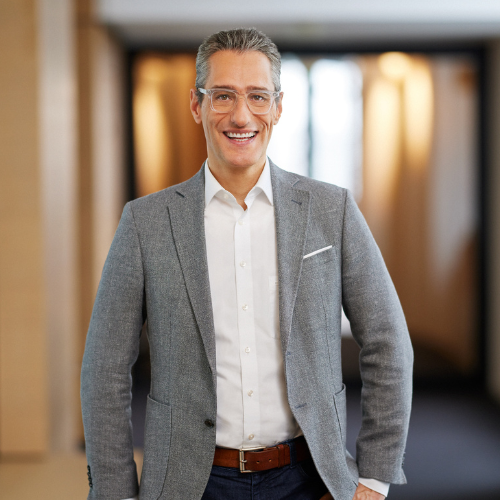 Eine Person im grauen Blazer lächelt in einem modernen, hellen Raum.