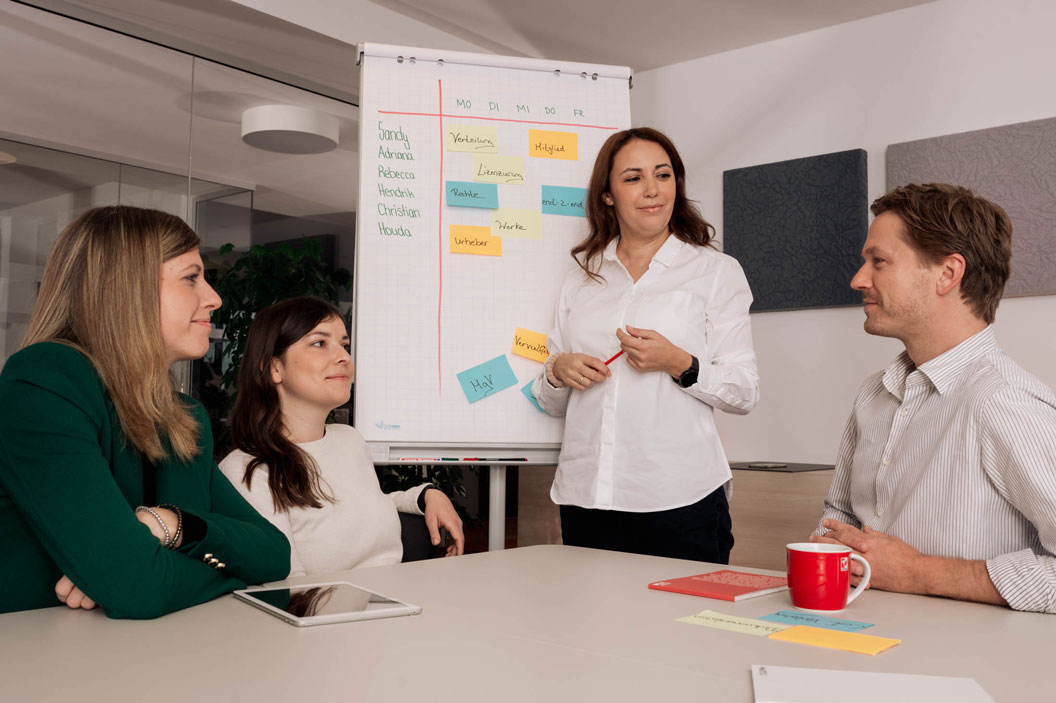 Vier Personen in einem Meeting, eine Frau steht vor einem Flipchart mit bunten Notizen.