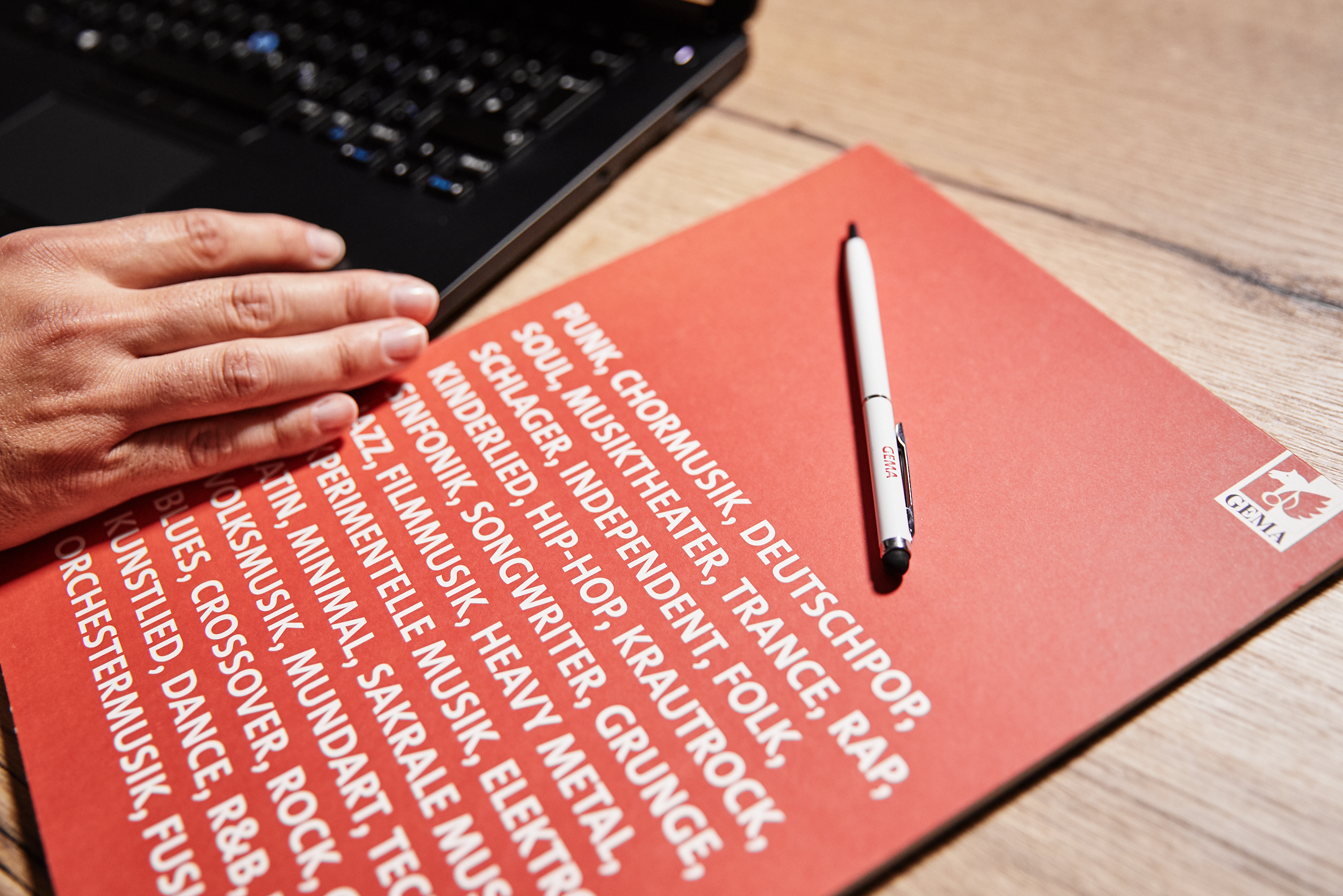 Hand on laptop, red paper with music genres, white pen on wooden table.