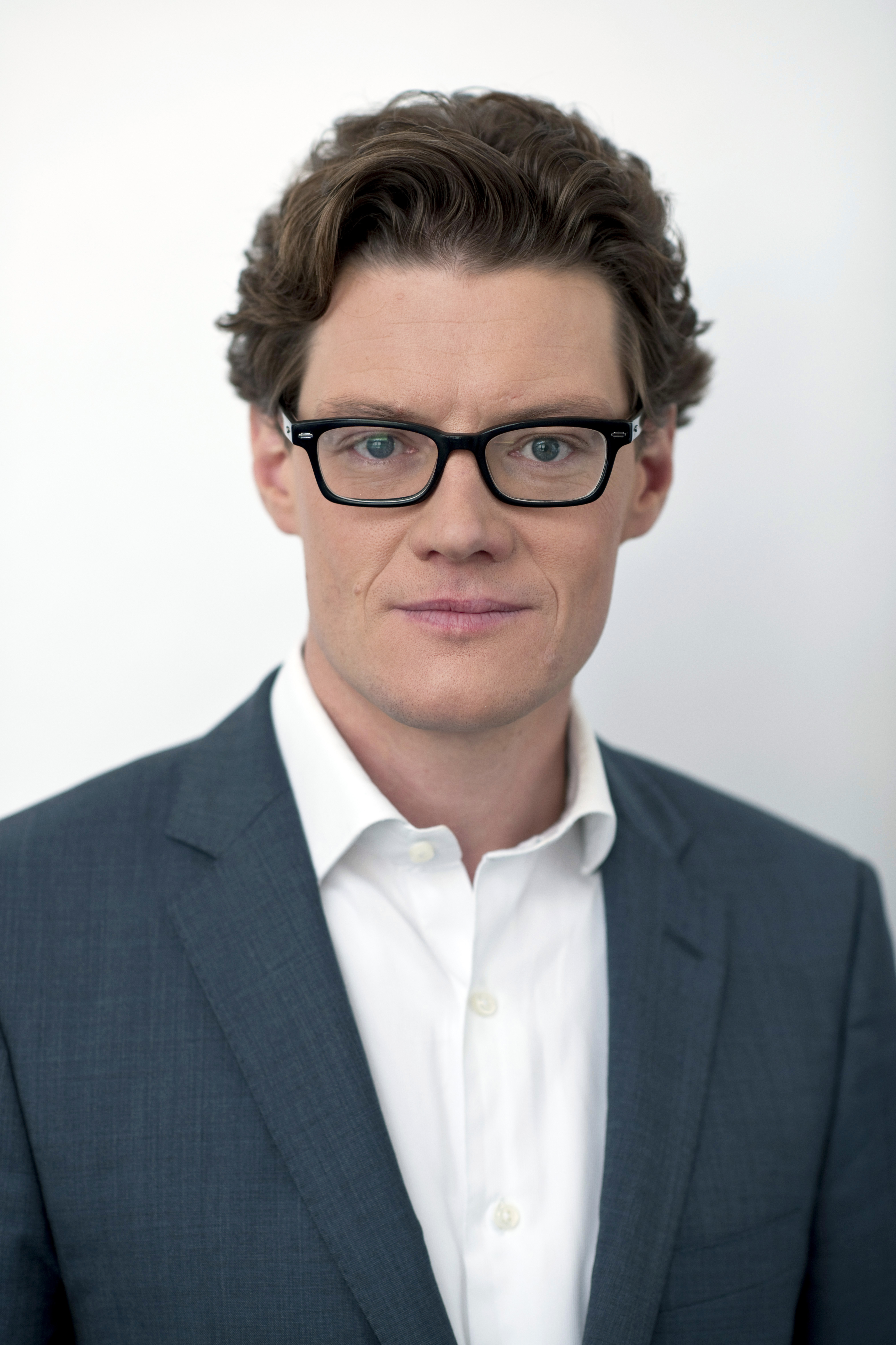Man with glasses in a gray suit and white shirt against a plain white background.