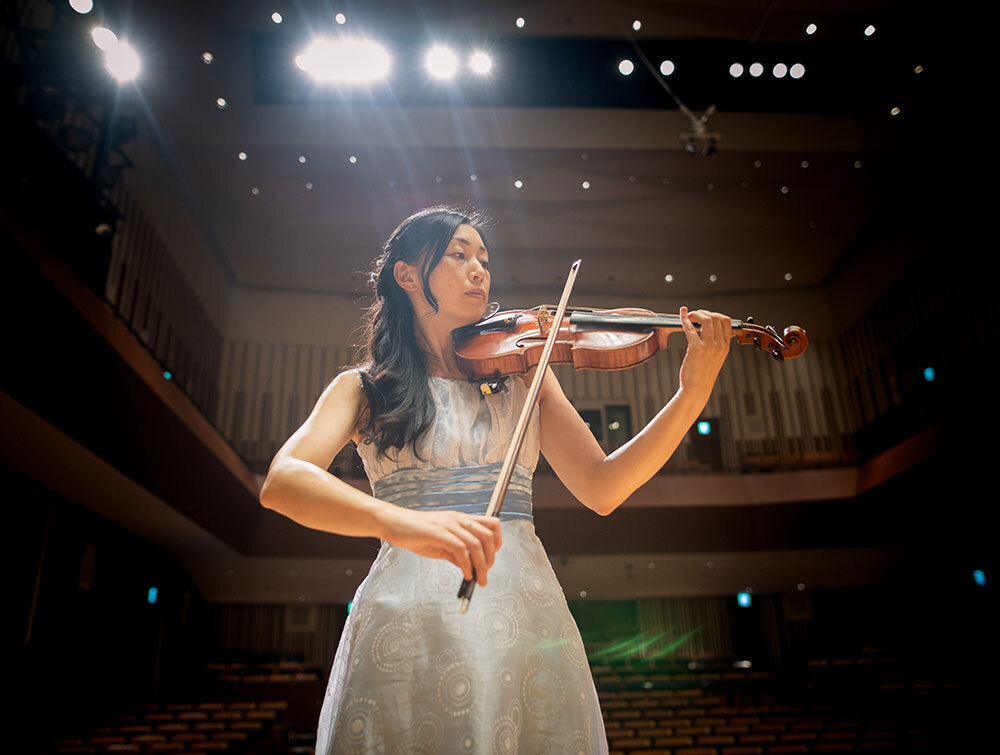 A person in a blue dress plays a violin on a stage under bright lights in a concert hall.