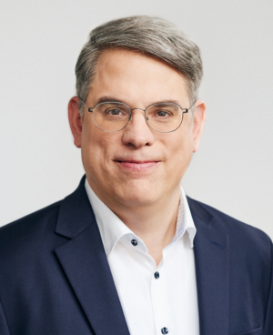 Eine Person mit Brille und grauem Haar in einem dunklen Anzug vor hellem Hintergrund.