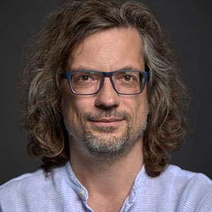 A person with long curly hair, wearing glasses and a light blue shirt, smiles slightly against a dark background.