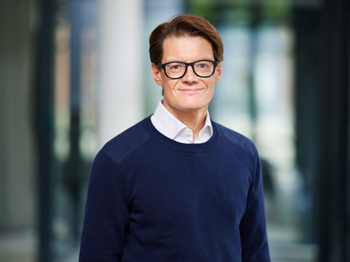 A person with glasses, wearing a navy sweater and white shirt, stands in front of a blurred indoor background.
