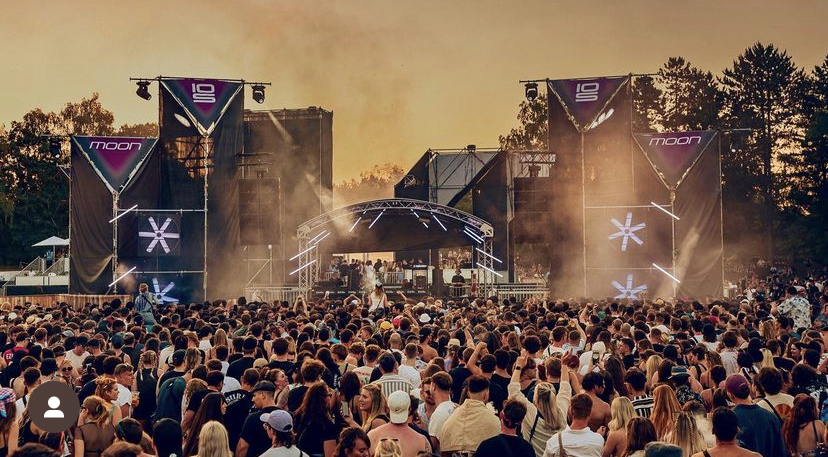 Menschenmenge vor einer Bühne bei einem Open-Air-Konzert bei Sonnenuntergang, umgeben von Bäumen.