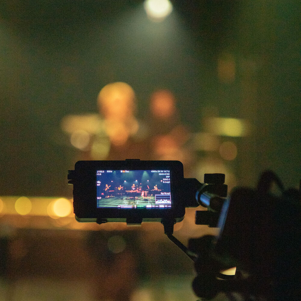 Camera screen shows a live concert stage with musicians, blurred audience and stage lights in the background.