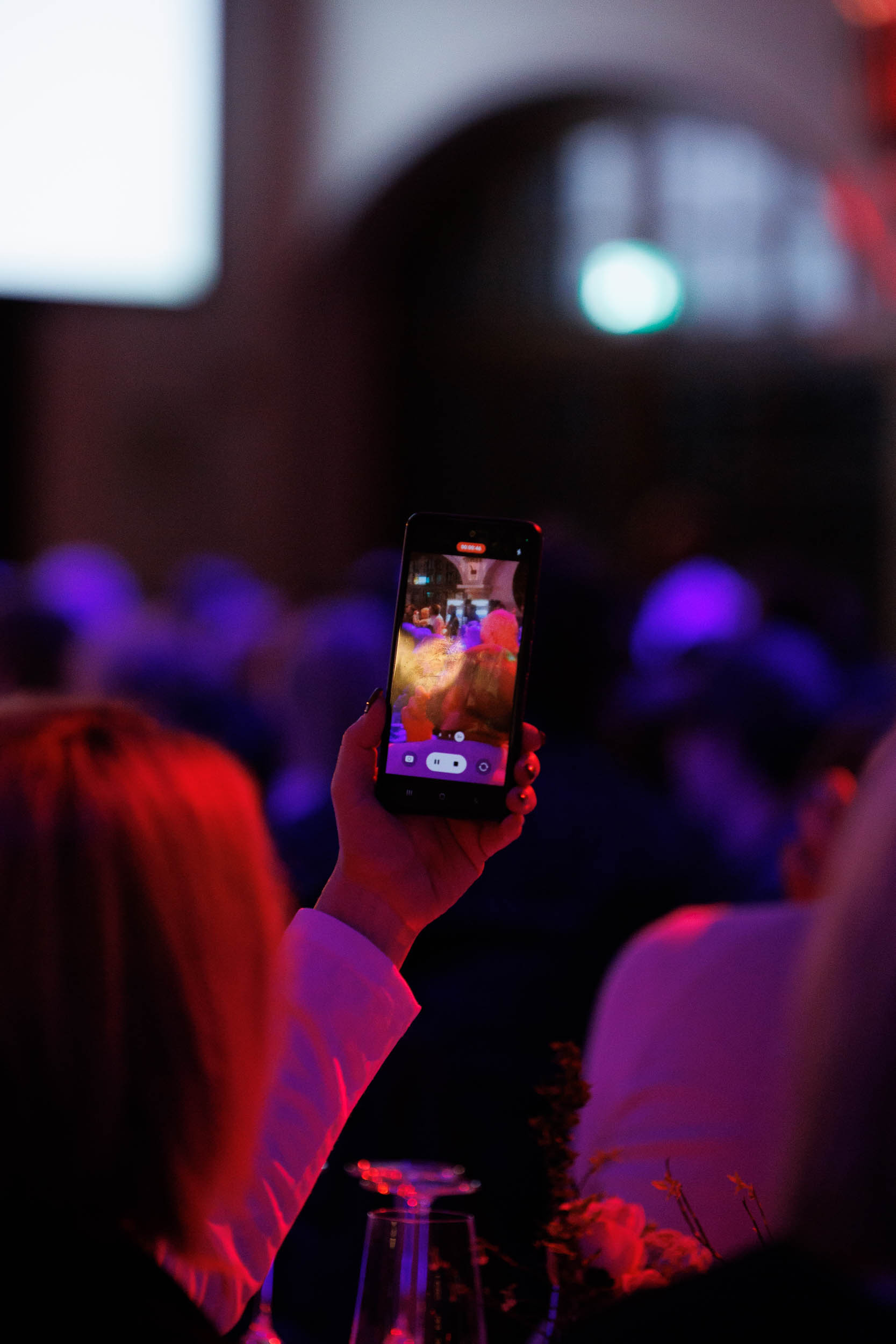 A person holds a smartphone recording a colorful event in a dimly lit room with red and blue lighting.