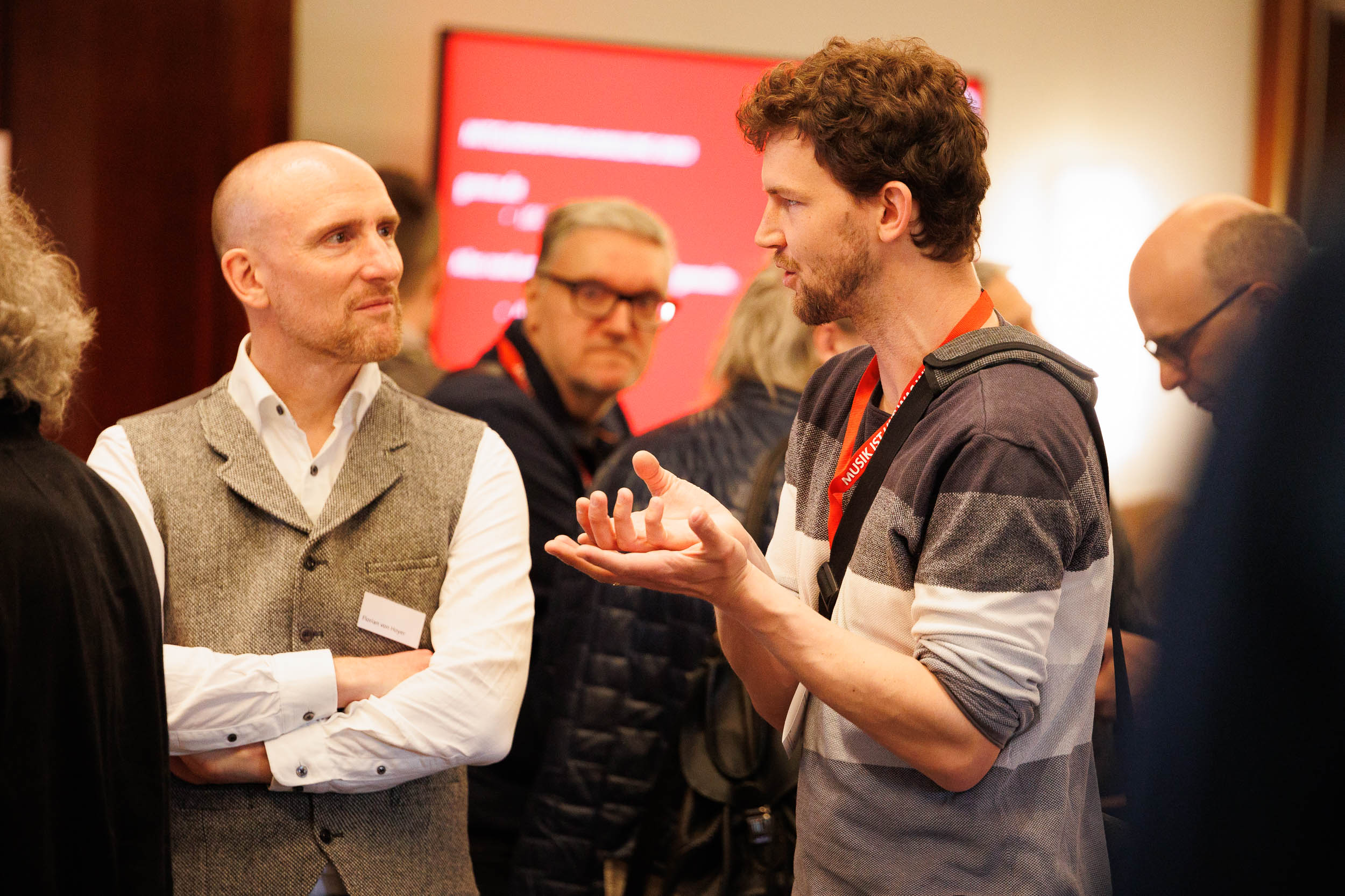 Two men engaged in conversation at an event, one gesturing with his hands, with people and a red screen in the background.