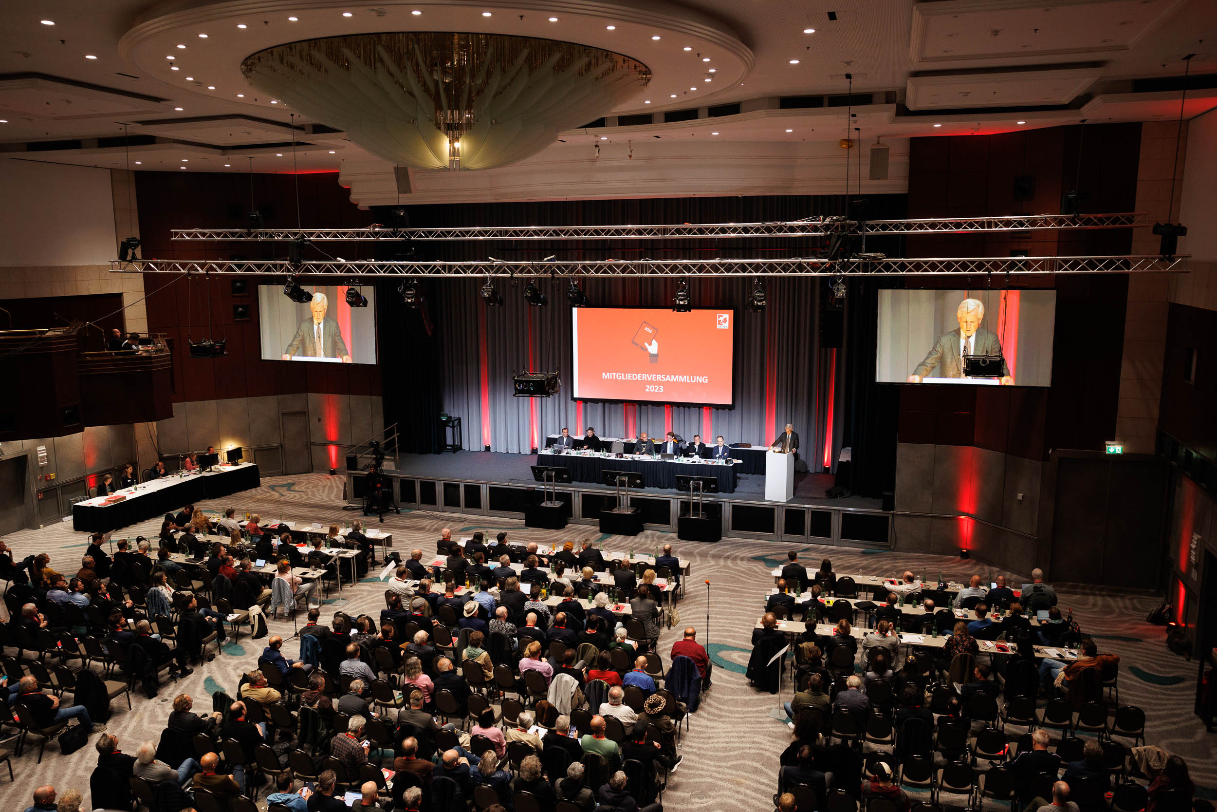 A conference hall with a speaker at a podium, audience seated, and a large screen displaying