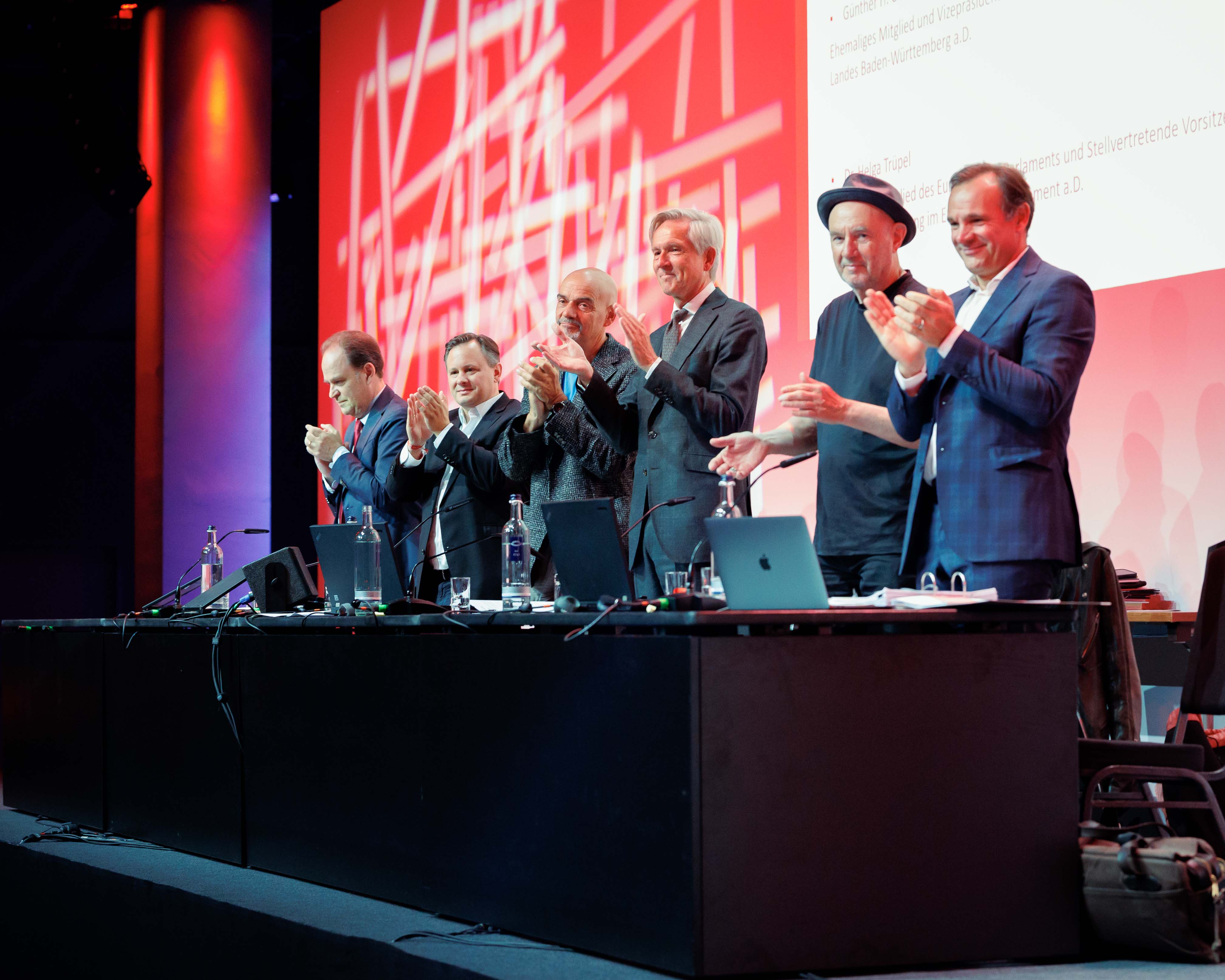 Six people stand behind a table clapping, with laptops and bottles, in front of a red and white backdrop.