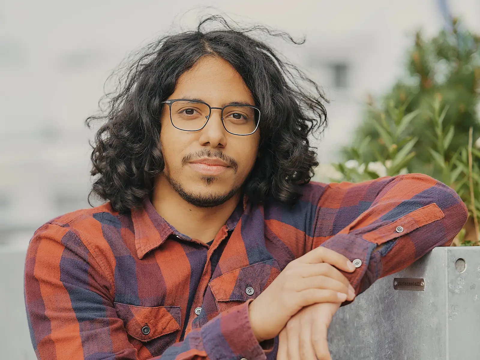 Eine Person mit Brille und lockigem Haar trägt ein kariertes Hemd und lehnt an einer Metallbarriere, im Hintergrund Pflanzen.