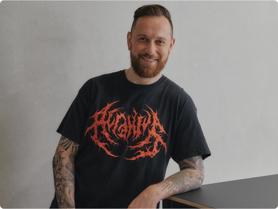 Ein Mann mit Bart lehnt lächelnd an einem Tisch, trägt ein schwarzes T-Shirt mit rotem Schriftzug.