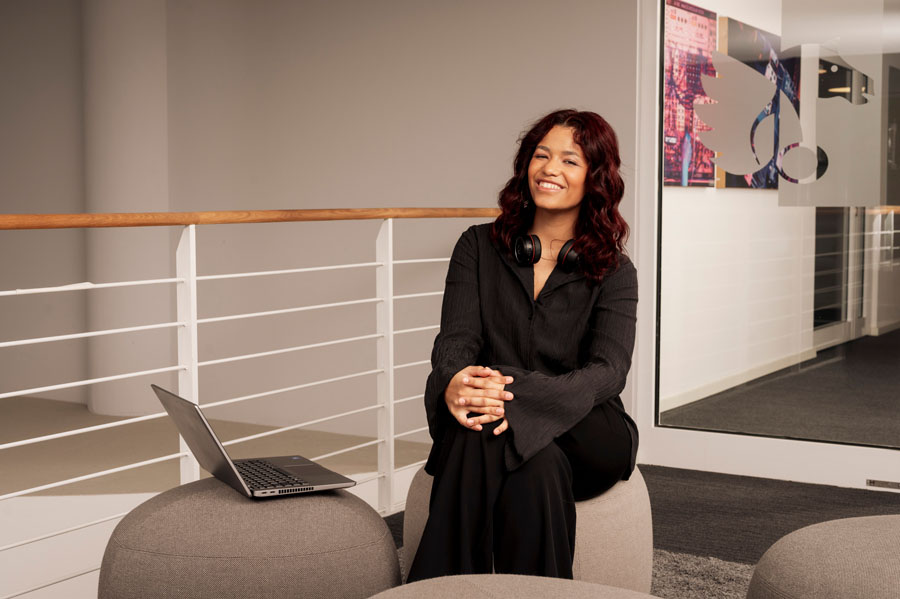 A woman in black sits on a gray ottoman, smiling, with a laptop nearby and headphones around her neck.