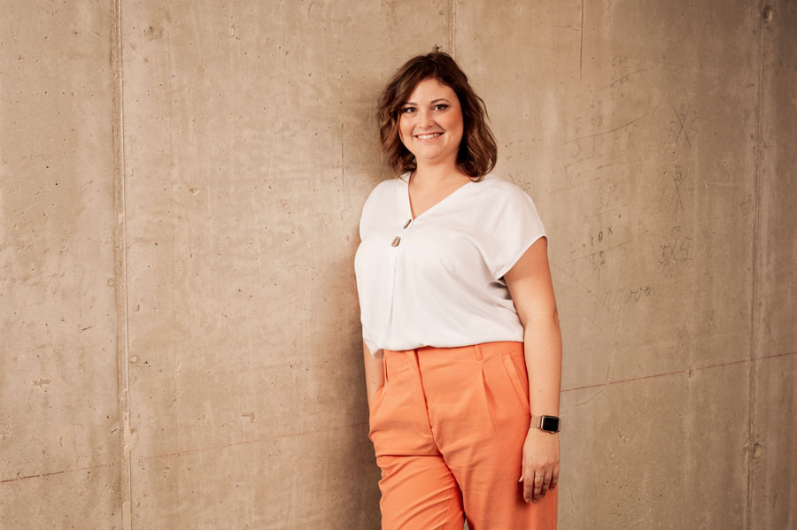 A person in a white blouse and orange pants smiles, leaning against a concrete wall.