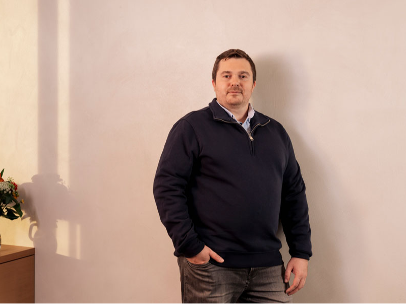 Ein Mann in dunkelblauem Pullover steht vor einer beigen Wand, links ein Blumenstrauß.