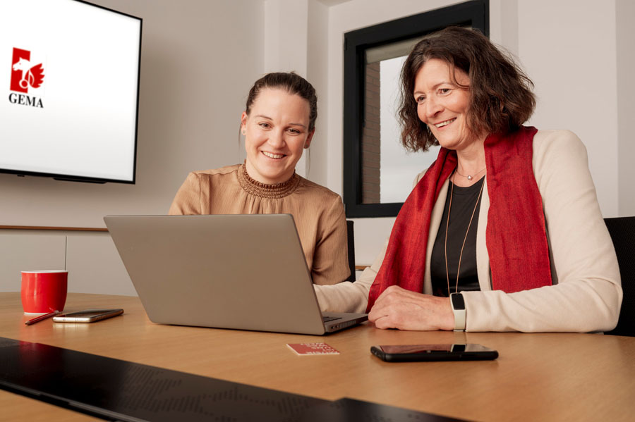 Zwei Frauen lächeln vor einem Laptop, roter Schal, rote Tasse, GEMA-Logo auf Bildschirm im Hintergrund.