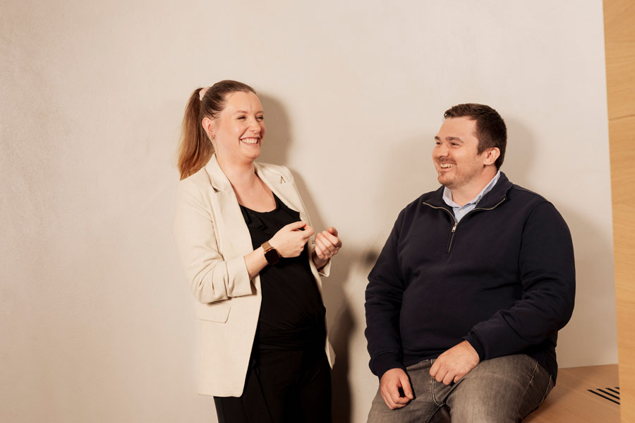 Eine Frau in einem beigen Blazer lacht mit einem Mann in einem dunklen Pullover vor einer hellen Wand.