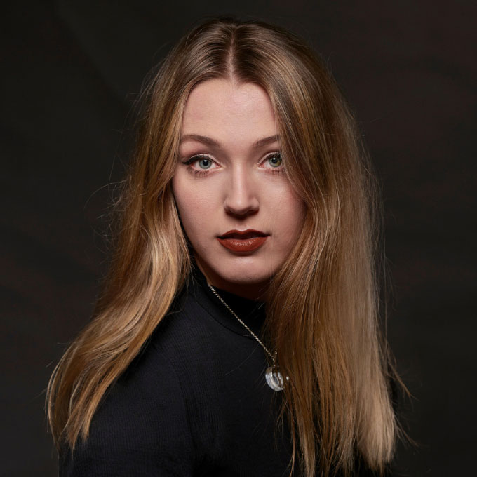 Eine Frau mit langen blonden Haaren trägt einen schwarzen Pullover vor dunklem Hintergrund.