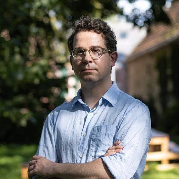Eine Person mit Brille und blauem Hemd steht mit verschränkten Armen im Garten.