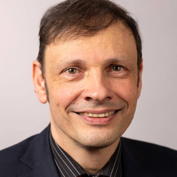 Lächelnder Mann in dunklem Anzug und gestreiftem Hemd, mit kurzem Haar, vor einem schlichten Hintergrund.