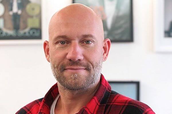 Eine bärtige Person in einem rotkarierten Hemd steht in einem Raum, hinter ihr hängen verschwommene gerahmte Bilder an der Wand.
