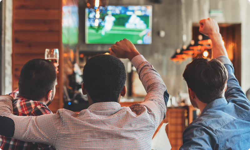 Three people cheer while watching a sports game on a TV in a cozy bar.