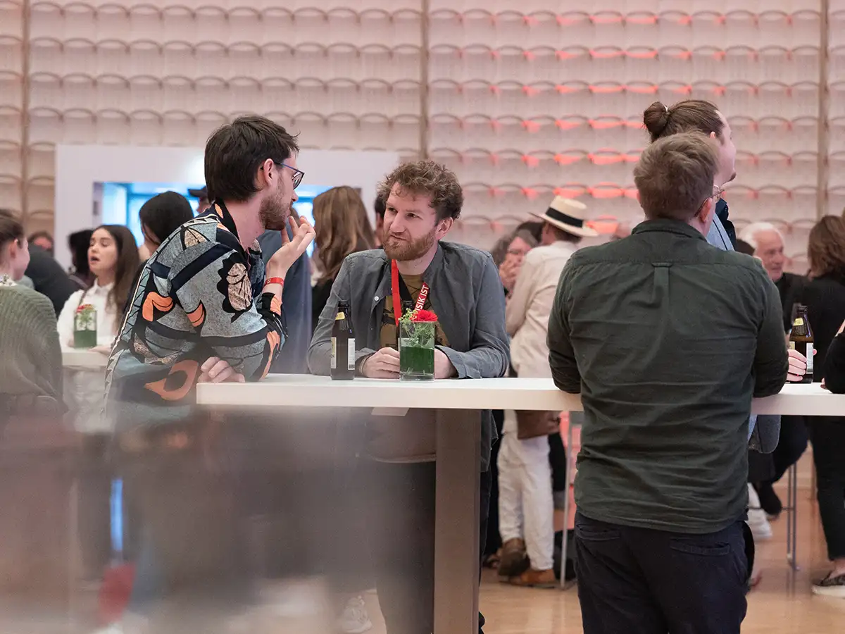 People socialize around a high table with drinks in a busy room, featuring a textured wall and warm lighting.