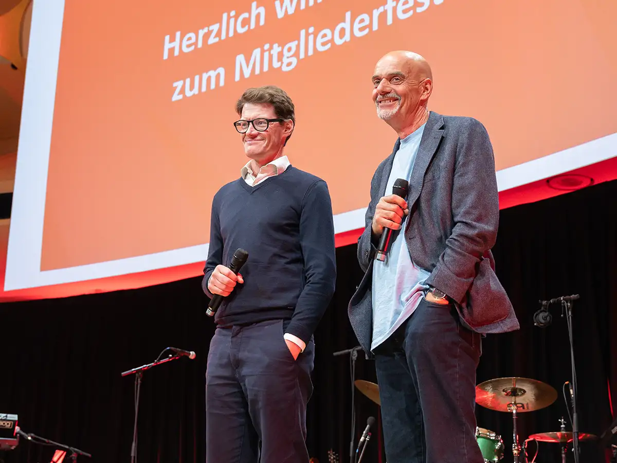 Two men with microphones stand on stage, smiling, in front of an orange screen with text.