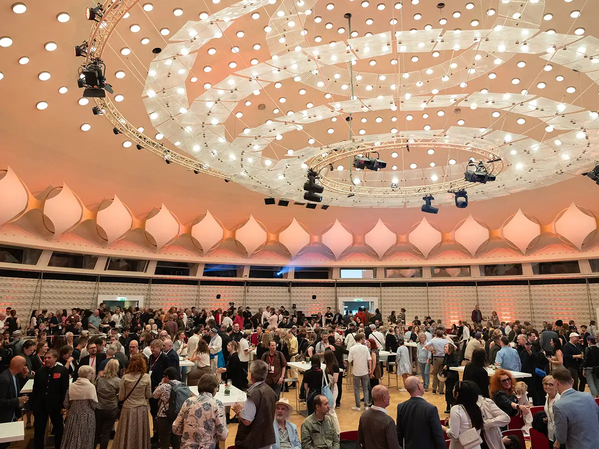 A large crowd gathers in a modern hall with a bright, circular ceiling and decorative lighting.
