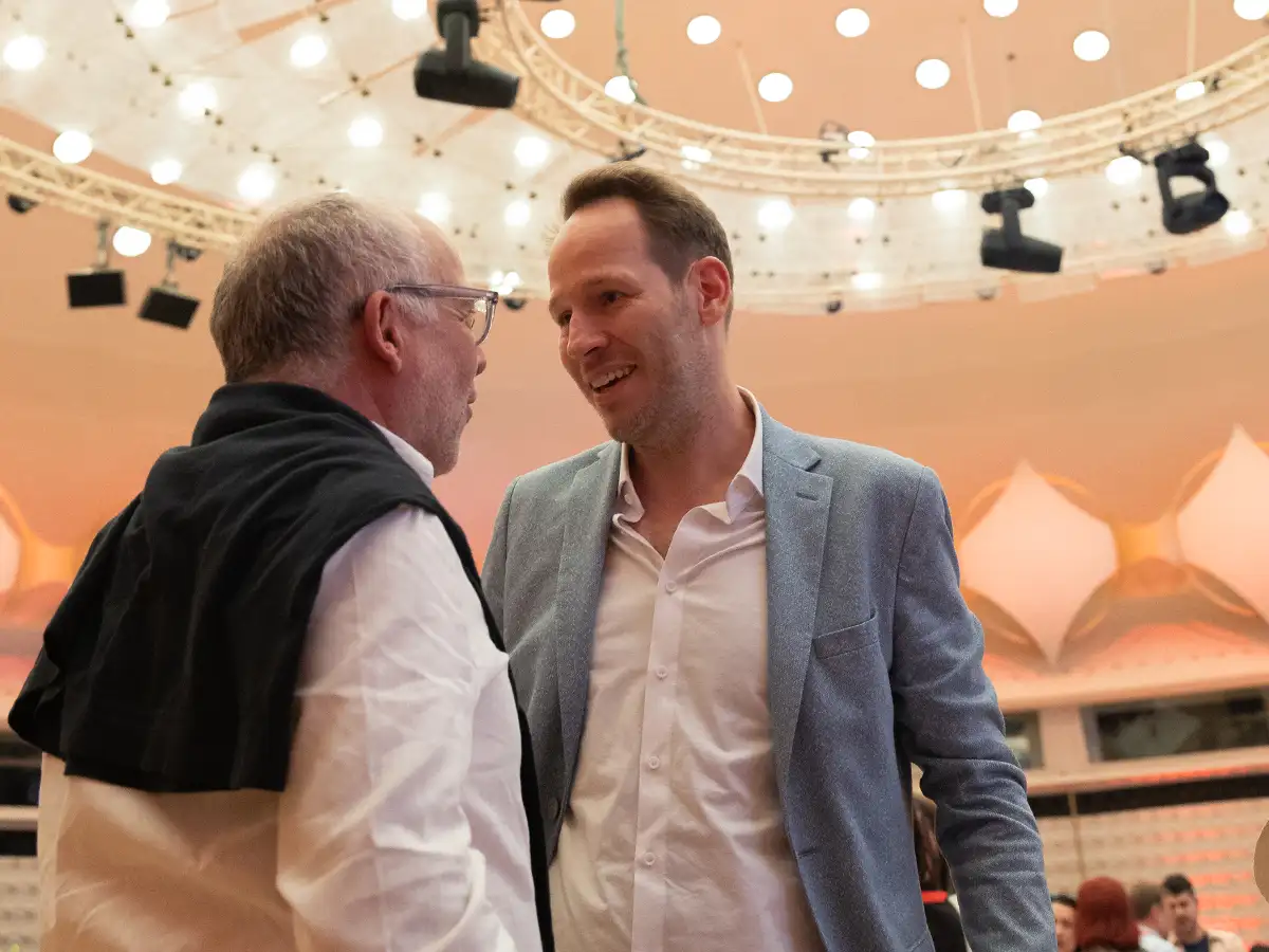 Two men in conversation under a ceiling with bright circular lights. One wears a jacket, the other a sweater over shoulders.