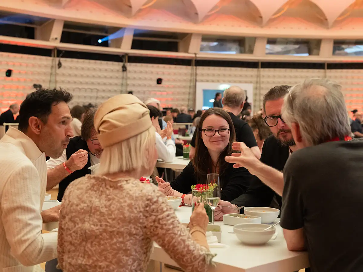 A group of people sit at a table in a large room, talking and eating, with drinks and flowers on the table.