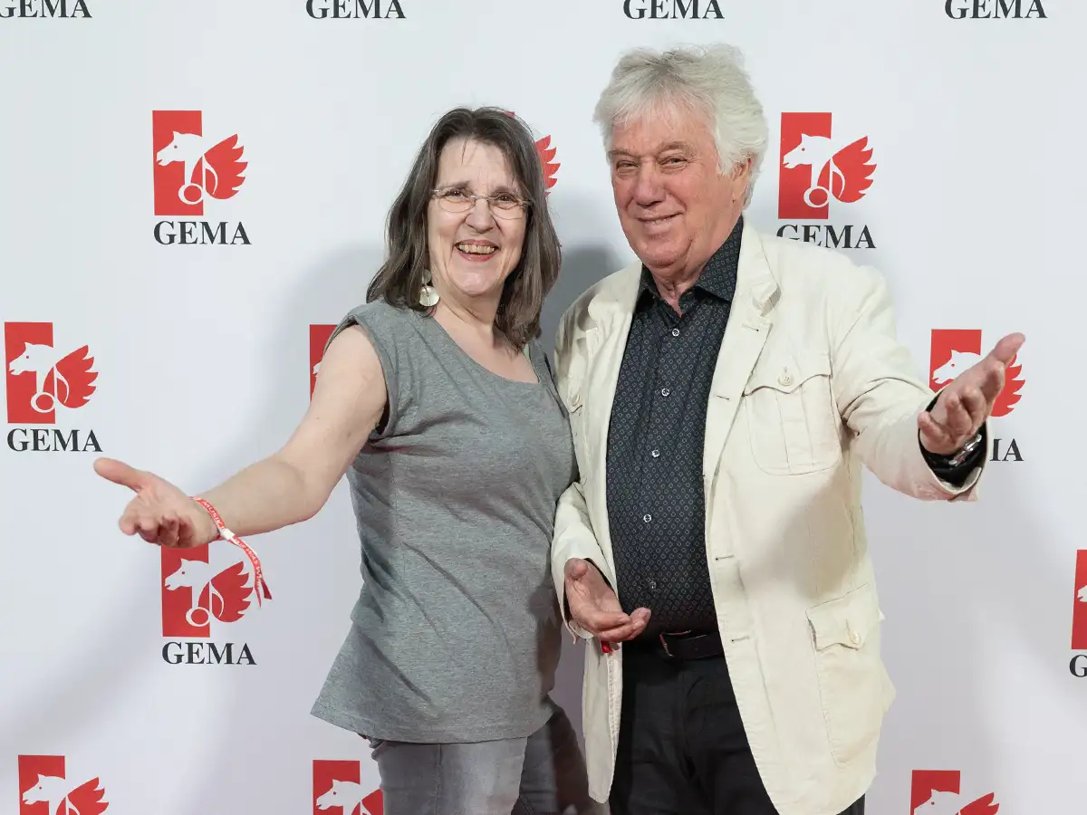 Two people smiling with open arms at a GEMA event, standing in front of a logo backdrop.