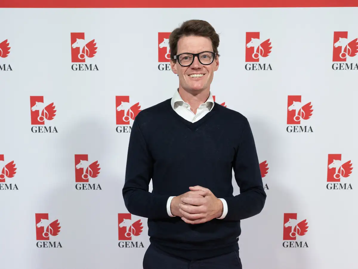A person in glasses and a dark sweater stands smiling in front of a GEMA logo backdrop.