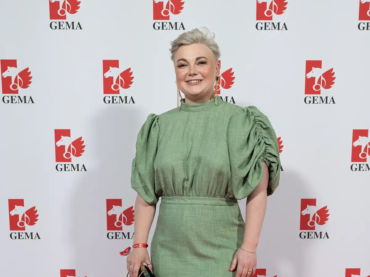 Person in a green dress smiles on a red carpet with GEMA logos in the background.