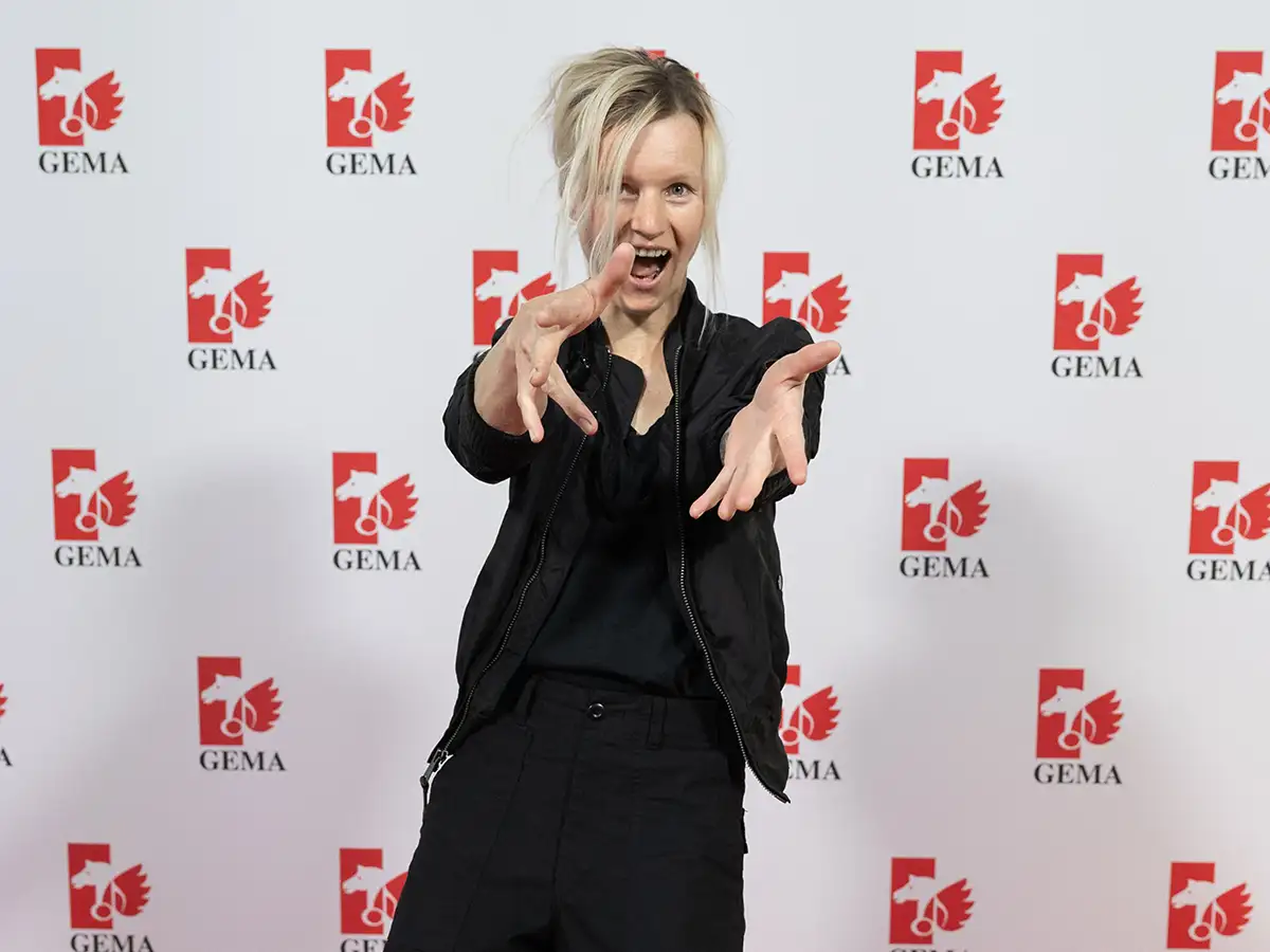 Person in black outfit poses energetically in front of a GEMA logo backdrop.