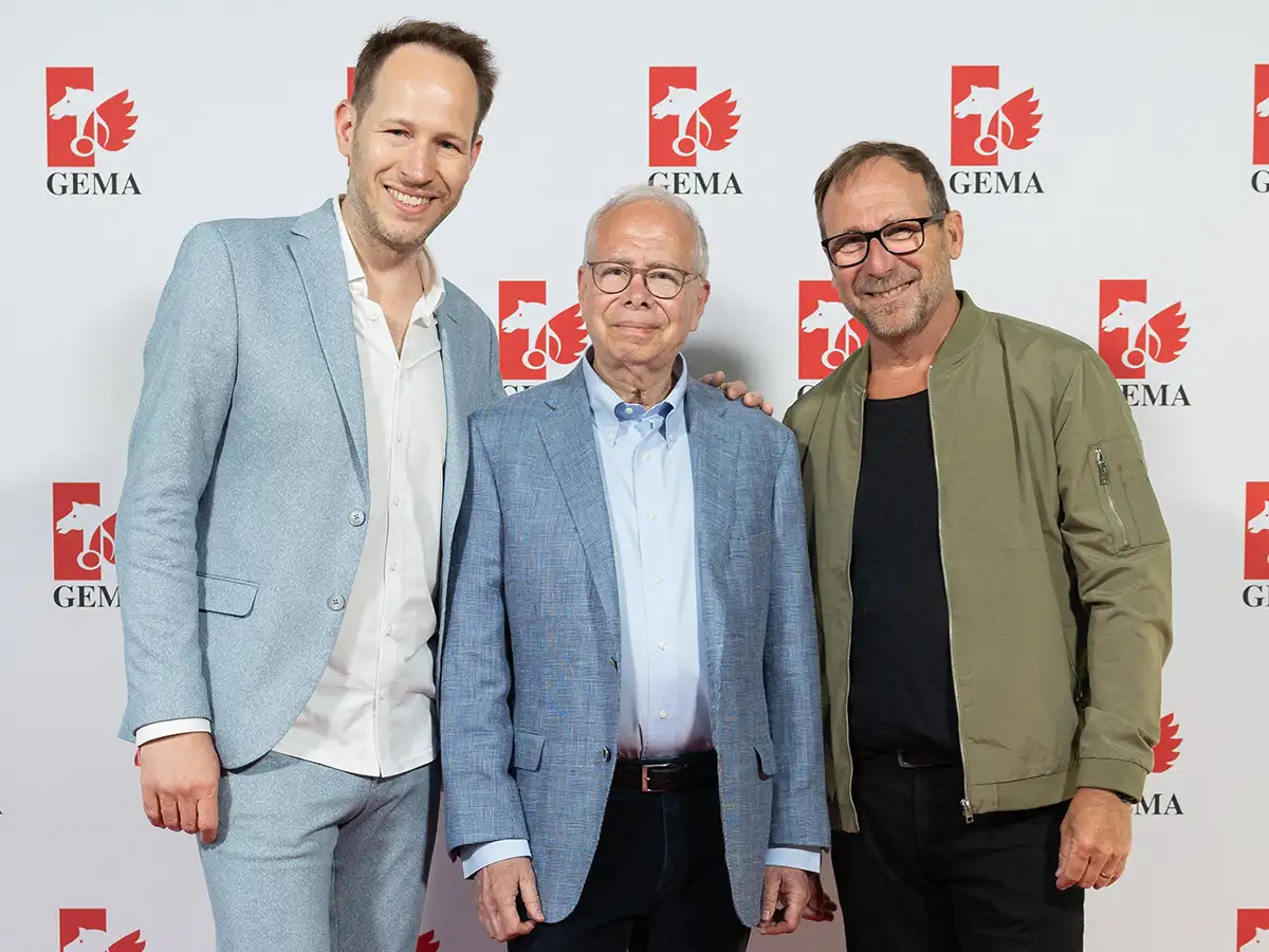 Three men in suits and jackets stand smiling in front of a GEMA logo backdrop.