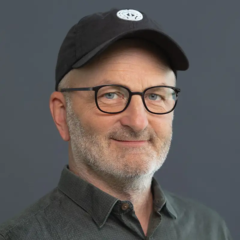 Ein Mann mit Brille und schwarzer Kappe lächelt vor grauem Hintergrund.