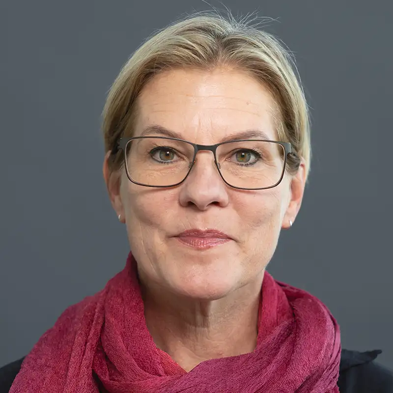 Eine Person mit Brille und rotem Schal vor grauem Hintergrund.
