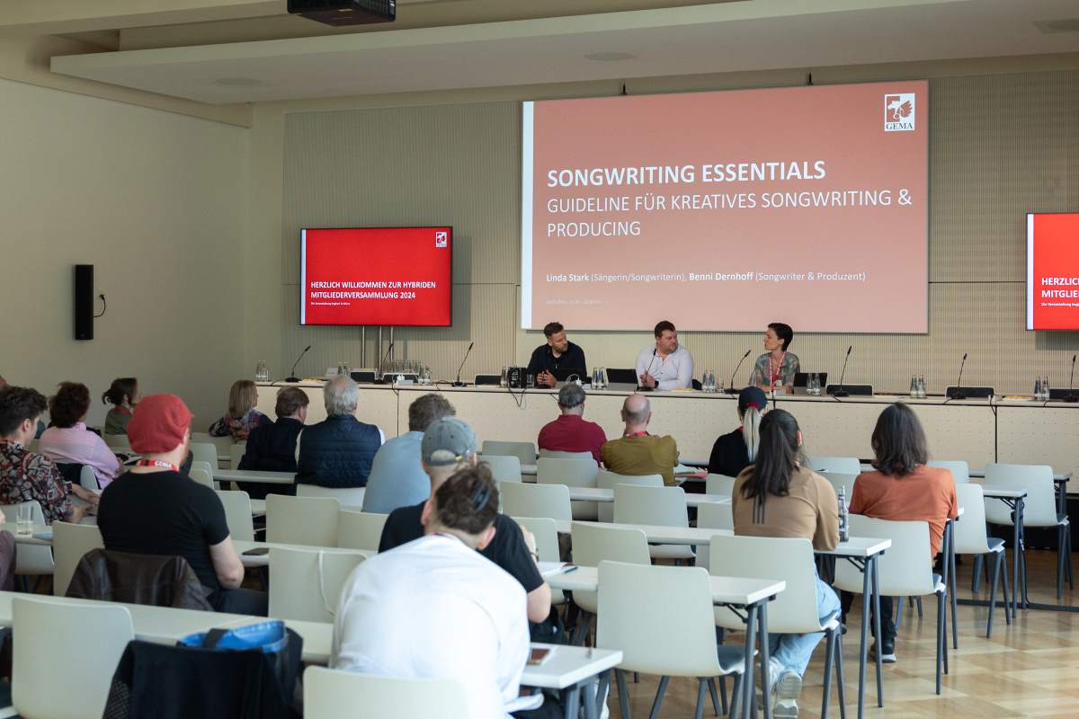 People sit in a conference room facing a panel discussing songwriting essentials, with red presentation screens at the front.