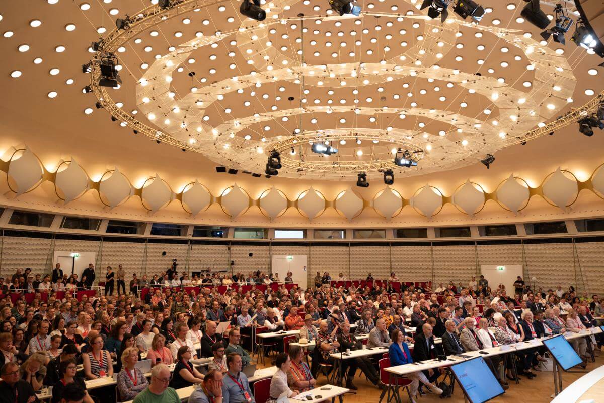 A large audience sits in a modern auditorium with circular ceiling lights and multiple screens at the front.