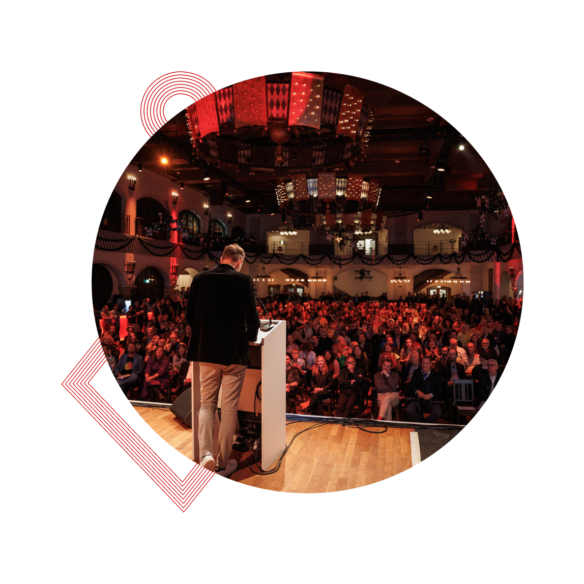 A speaker at a podium addresses a large seated audience in a warmly lit auditorium with chandeliers.