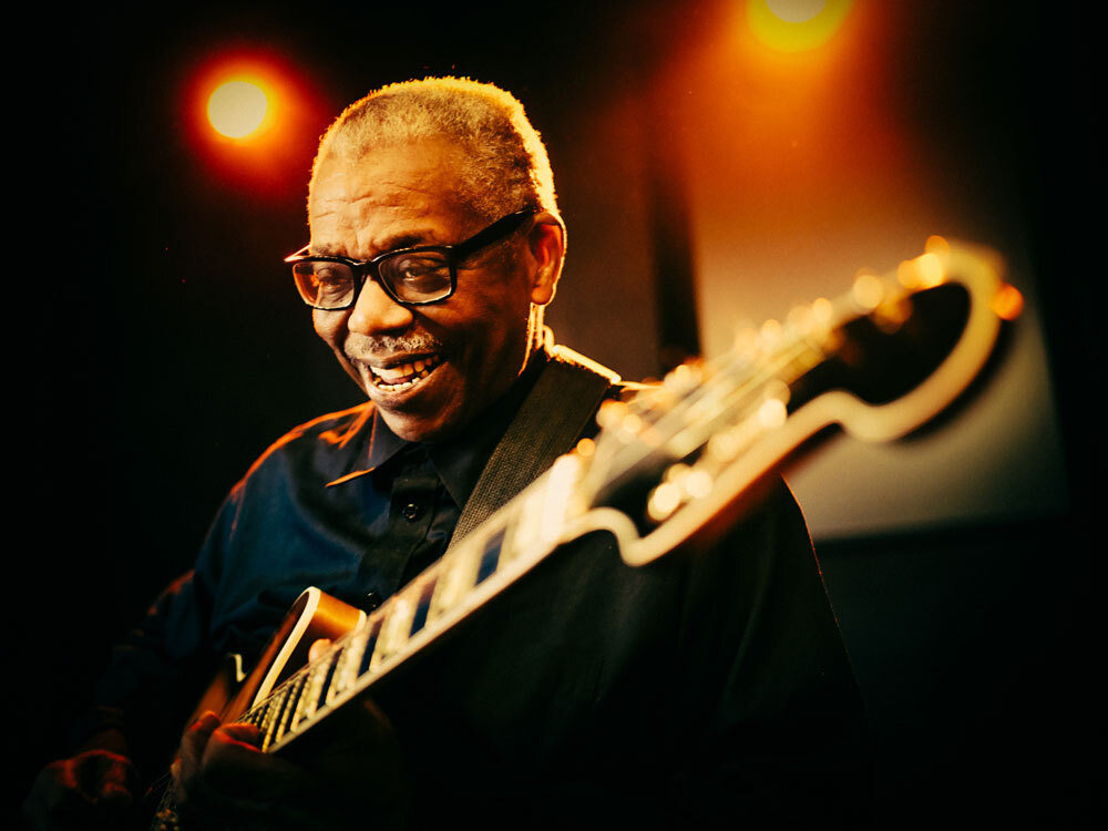 An elderly man in glasses smiles while playing an electric guitar under warm stage lights.
