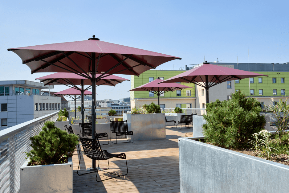 A rooftop terrace with red umbrellas, black chairs, and green plants under a clear blue sky.