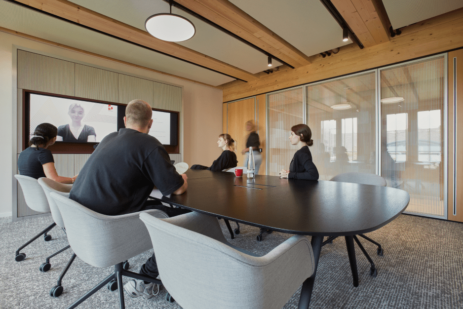 Five people in a modern conference room, watching a video call on a large screen, with wooden accents and glass walls.